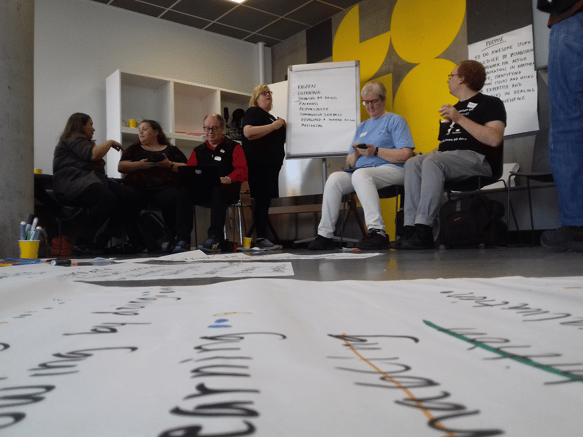 Group of people sitting in a workshop setting with papers laid out on the floor and a whiteboard with notes displayed.