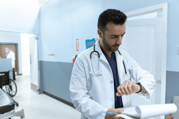 A male doctor in a white coat and stethoscope checking his watch while standing in a hospital hallway.