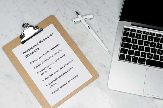 A clipboard with a COVID-19 protective measures checklist on a marble desk next to a laptop, pen, and paper clips.