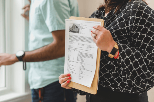 A person holding a document with a picture of a house and other information on it, with another person in a light blue shirt in the background.&nbsp;