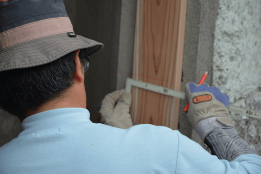 Man in blue shirt and brown hat inspecting a building using a ruler