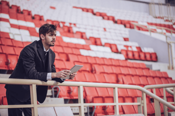 Man in a suit holding a tablet while leaning on a railing in an empty hockey stadium with red and white seats, symbolizing team management and planning.